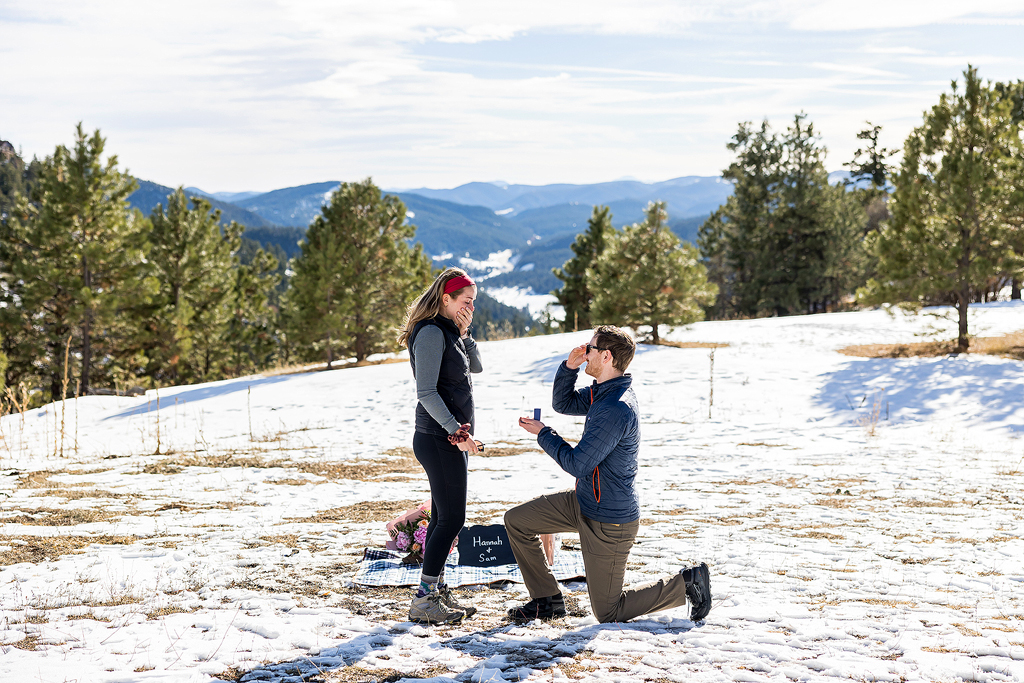 Sam kneels in a snowy Colorado meadow to propose to Hannah at Mt Falcon.