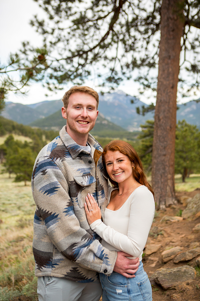 Noah and Lea smile under a large pine tree after their Colorado engagement.