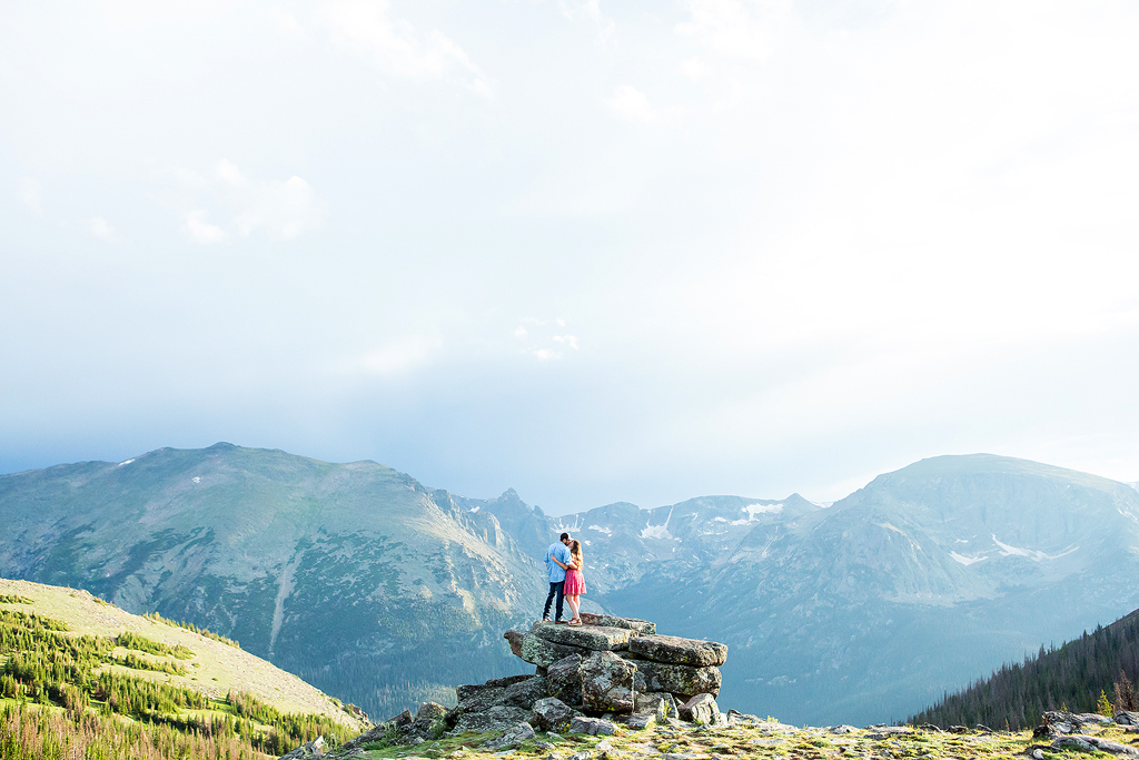 Zach and Sabrina kiss on a rock outcropping with a panoramic mountain view behind.