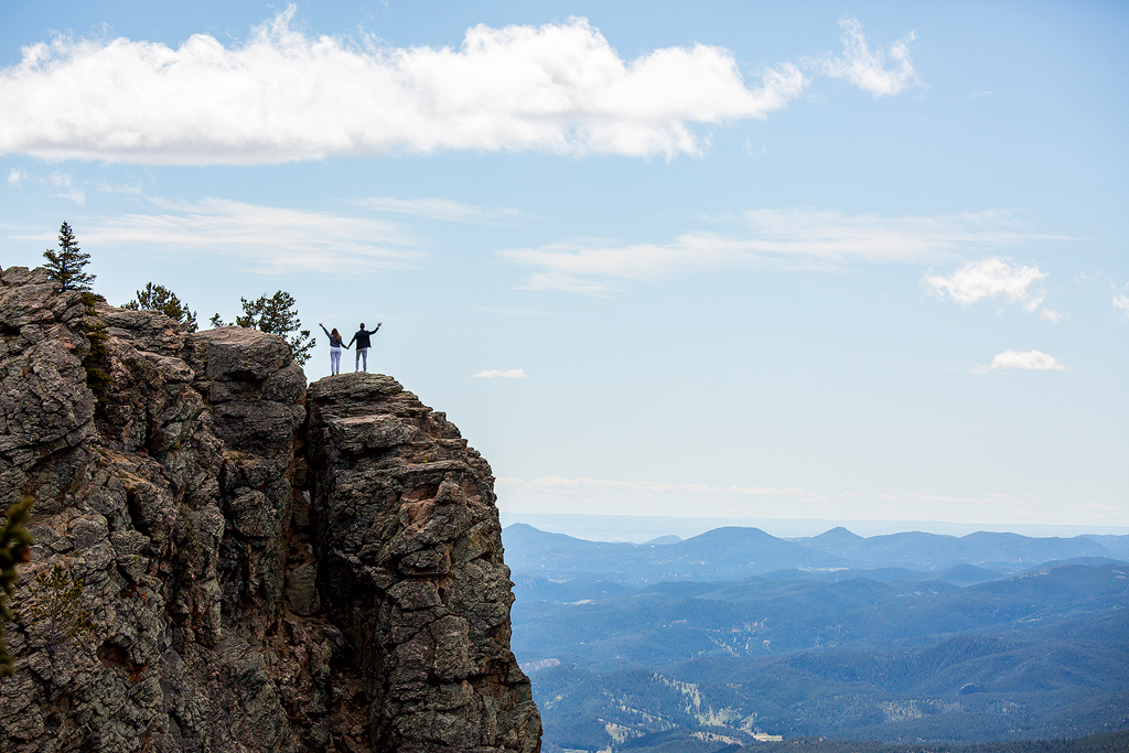 Couple stands on top of rocky cliffs with mountain views in the distance at Juniper Pass in Colorado.